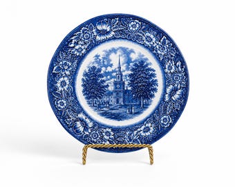 May include: A blue and white decorative plate featuring a detailed church scene in the center. The plate has a floral border and is displayed on a gold-colored stand. The plate's design is reminiscent of classic blue and white china.