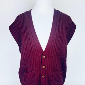 May include: A burgundy knitted sleeveless sweater vest with a V-neck and wooden buttons. The vest has two pockets on the front.