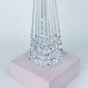 May include: A clear crystal decorative object with a geometric design. The base is a series of stacked cubes, and the top is a tall, faceted pyramid shape. It sits on a light pink rectangular block.