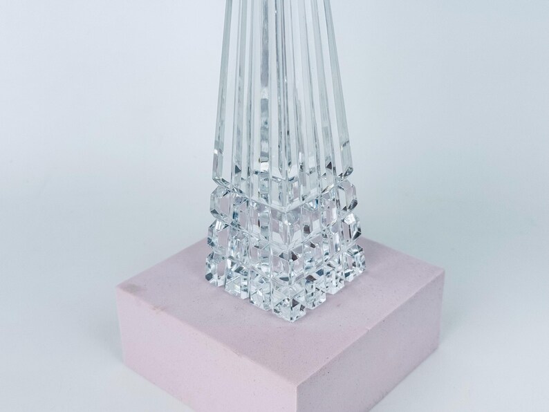 May include: A clear crystal decorative object with a geometric design. The base is a series of stacked cubes, and the top is a tall, faceted pyramid shape. It sits on a light pink rectangular block.