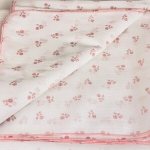May include: A white muslin baby blanket with a pink floral pattern. The blanket is folded in half and has a pink trim.