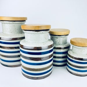 May include: Set of four ceramic canisters with wooden lids. The canisters are white with blue and brown stripes.
