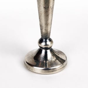 May include: A silver-toned metal candlestick base featuring a smooth, tapered stem that rises from a rounded, bulbous connector piece, which sits atop a wide, circular, flat-bottomed pedestal stand with a polished, reflective metallic finish.