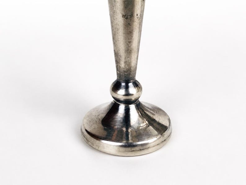 May include: A silver-toned metal candlestick base featuring a smooth, tapered stem that rises from a rounded, bulbous connector piece, which sits atop a wide, circular, flat-bottomed pedestal stand with a polished, reflective metallic finish.