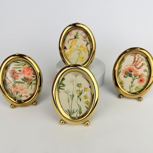May include: Four oval gold picture frames, each with a floral print. The frames stand on small feet. The prints display flowers in orange, yellow, and green on a cream background. Decorative home decor, each frame is approximately 7 cm tall.