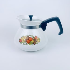 Könnte beinhalten: Weiße Emaille-Teekanne mit schwarzem Griff und Deckel. Die Teekanne hat ein farbenfrohes Blumen- und Fruchtdesign um den Boden. Ein silbernes Band umschließt die Mitte der Teekanne.