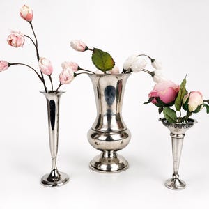 May include: Three silver-toned metal vases of varying shapes and sizes. The left vase is tall and slender, the center vase is wide with a flared rim, and the right vase is shorter with a scalloped edge. Each contains artificial pink and white flowers.