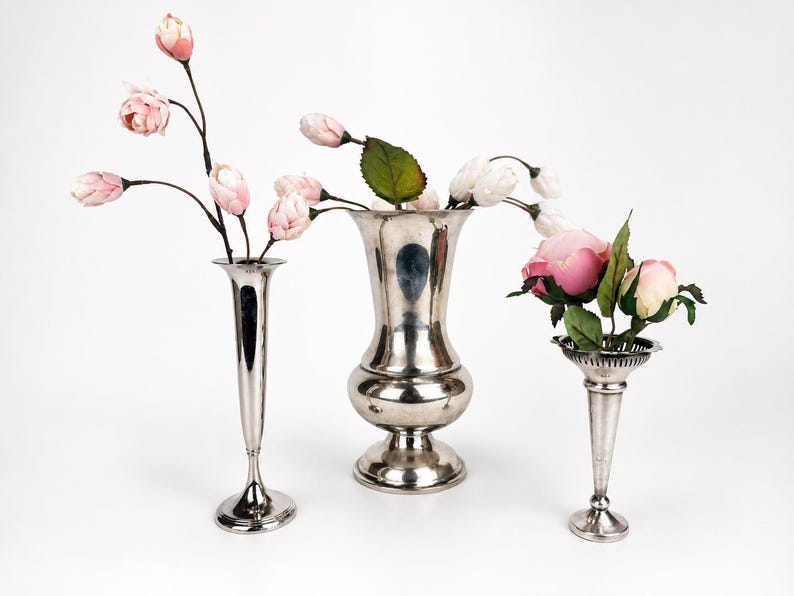 May include: Three silver-toned metal vases of varying shapes and sizes. The left vase is tall and slender, the center vase is wide with a flared rim, and the right vase is shorter with a scalloped edge. Each contains artificial pink and white flowers.