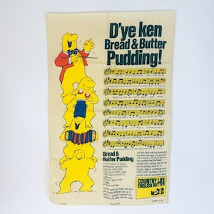 May include: Cream-colored kitchen towel with the text "D'ye ken Bread & Butter Pudding!" and a recipe. The towel features a stack of yellow cartoon figures, musical notes, and a recipe for bread and butter pudding.