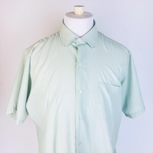 May include: A light green short-sleeved button-down shirt with a chest pocket. The shirt is displayed on a white mannequin. The shirt has a classic collar and buttoned cuffs. The fabric appears to be a lightweight cotton or blend.