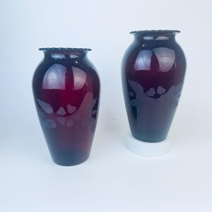 May include: Two dark red glass vases with etched floral designs. The vases are similar in shape and size, with a slightly wider base and a narrow neck.