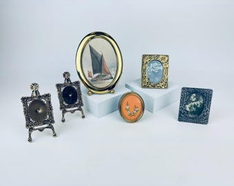 May include: Five ornate picture frames with different designs and colors. The frames are made of metal and have a vintage look. Some of the frames are on small stands.