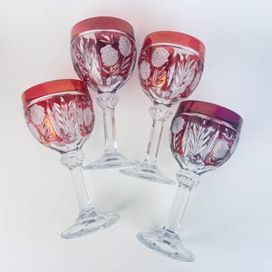 May include: Four crystal wine glasses with ruby red rims and floral etched designs. The clear stems and bases are intricately cut. The glasses are arranged on a white surface, showcasing their elegant details and craftsmanship.