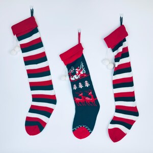 Puede incluir: Tres calcetines de Navidad colgados en una pared blanca. Los calcetines son de rayas rojas y blancas, de rayas rojas y blancas con un diseño de reno, y azules con un diseño de reno.