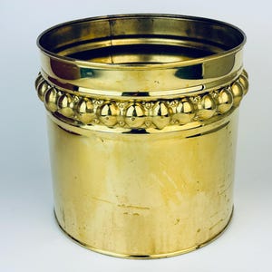 May include: A gold metal planter with a beaded rim. The planter is cylindrical and has a smooth, shiny surface.