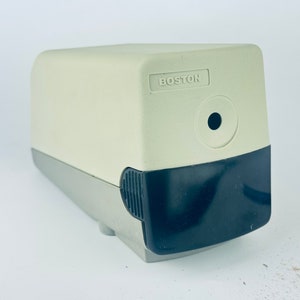 May include: A cream-colored Boston pencil sharpener with a black front and a gray base. The word "BOSTON" is printed on the top. A pencil and shavings are visible in the background.