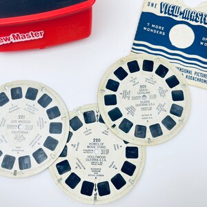 Puede incluir: Un visor View-Master rojo con tres carretes antiguos con imágenes de Los Ángeles, Santa Catalina y Hollywood. Los carretes están etiquetados con ubicaciones y texto. También hay una tarjeta de carrete View-Master azul y blanca.