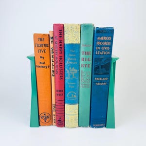 Pode incluir: Um conjunto de sete livros antigos de capa dura com capas coloridas, exibidos em um suporte de livros turquesa. Os títulos incluem "The Fighting Five", "My Spelling", "The Happy Hollisters", "The Big Eye", e "America's Progress in Civilization".