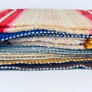 May include: A stack of rectangular fabric swatches featuring various textures and colors. The top layer is a beige and red striped textile, followed by layers of blue, white, grey, tan, and brown woven materials with distinct edge finishes.