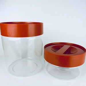 Puede incluir: Dos tarros de almacenamiento de vidrio transparente con tapas marrones. Uno es alto y cilíndrico, el otro más corto y ovalado. Las tapas tienen un diseño plano con un asa empotrada. Los tarros están vacíos.