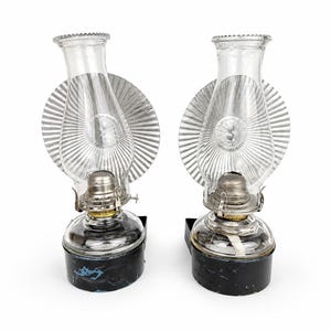 May include: A pair of wall-mounted oil lamps featuring clear glass chimneys, silver-toned metal reflectors with a radial ribbed pattern, and black circular bases. Each lamp includes a metallic burner assembly and a visible wick inside the glass.