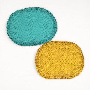 May include: Two oval-shaped placemats with ruffled edges. One placemat is teal with a quilted wavy pattern, and the other is mustard yellow with a quilted wavy pattern and small embroidered dots across the surface of the fabric.
