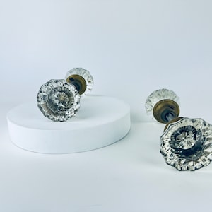 Peut inclure: Quatre poignées de porte en verre transparent avec un motif en spirale et des ferrures en laiton. Deux poignées sont sur un socle blanc, les autres à droite. Les poignées ont une esthétique vintage et antique.