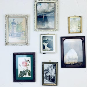 May include: A collection of framed art pieces, including landscape paintings, botanical prints, and architectural sketches, displayed on a white wall. The frames vary in style, from ornate gold to simple dark wood, creating a vintage aesthetic.