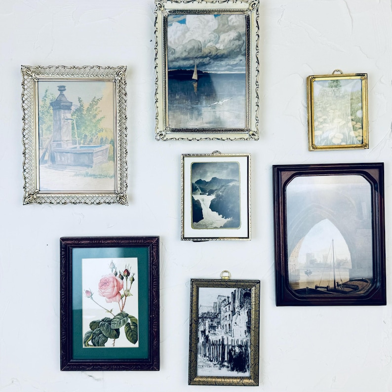 May include: A collection of framed art pieces, including landscape paintings, botanical prints, and architectural sketches, displayed on a white wall. The frames vary in style, from ornate gold to simple dark wood, creating a vintage aesthetic.