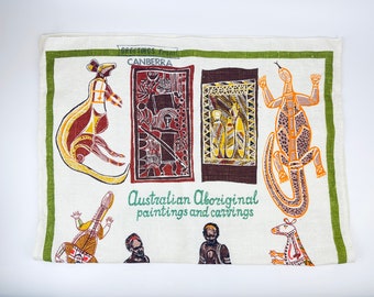 Op de afbeelding: Een witte theedoek met een groene rand heeft een kleurrijk ontwerp met Australische Aboriginal schilderingen en houtsnijwerk. Het ontwerp bevat een kangoeroe, een hagedis en verschillende figuren. De tekst "Greetings from Canberra" en "Australian Aboriginal paintings and carvings" staat op de theedoek gedrukt.