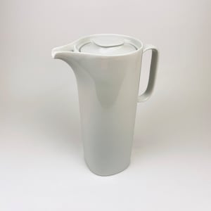 May include: A white ceramic pitcher with a lid and a handle. The pitcher is tall and slender, with a rounded base and a slightly flared top.