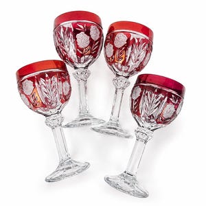 May include: A set of four crystal wine glasses featuring a deep red overlay at the rim and bowl. Each glass is decorated with intricate cut-glass floral patterns and leaf motifs, supported by a clear faceted stem and a circular base.