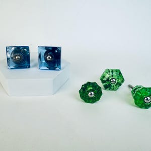 May include: An assortment of decorative cabinet knobs in shades of blue and green. Two square, faceted blue knobs with silver accents sit on a white platform. Three green, faceted knobs are also displayed.