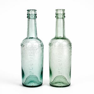 May include: Two vintage light green glass bottles with a slender neck and a rounded body. Each bottle features embossed text reading PERRIER on the front and a distinct indented base. The glass has a subtle aqua tint and a smooth, reflective surface.