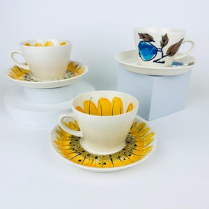 Peut inclure: Trois tasses à thé et soucoupes en céramique. Deux tasses ont des motifs floraux jaunes, et une a un motif de fruit bleu et de feuilles marron. Les tasses et les soucoupes sont blanches avec des accents jaunes et noirs.