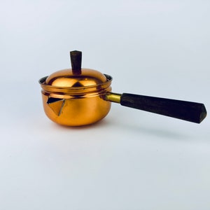 May include: A small copper-colored saucepan with a lid and a long, dark wooden handle. The handle is attached with a brass fitting. The saucepan has a geometric design on the side. The lid has a small wooden knob.