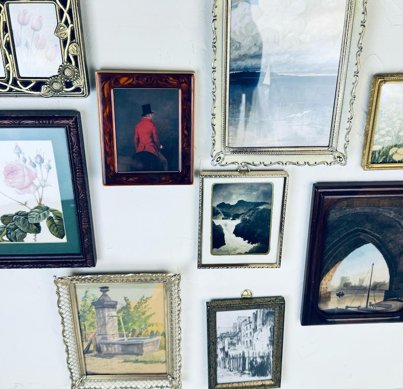 May include: Collection of framed art pieces, including a portrait of a person in a red coat, a landscape, and floral prints. Frames vary in style, from ornate gold to simple wood, creating a gallery wall aesthetic.