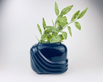May include: A navy blue ceramic vase with a glossy finish, featuring a wave-like design on the sides. The vase holds green leafy stems, creating a decorative arrangement. The vase is approximately 6 inches tall.