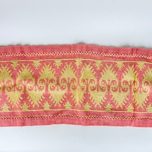 May include: A pink linen table runner with a yellow embroidered design. The design features a repeating pattern of stylized flowers and the word "Greetings".