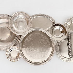 May include: A collection of silver-toned metal serving trays and bowls of various shapes and sizes, including round, oval, and rectangular pieces. Several items feature intricate embossed patterns, scalloped edges, and decorative borders.