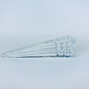 May include: A clear, glass door stop with a geometric, textured end and a tapered, ridged body. The door stop is lying horizontally on a white surface. The design is modern and minimalist.