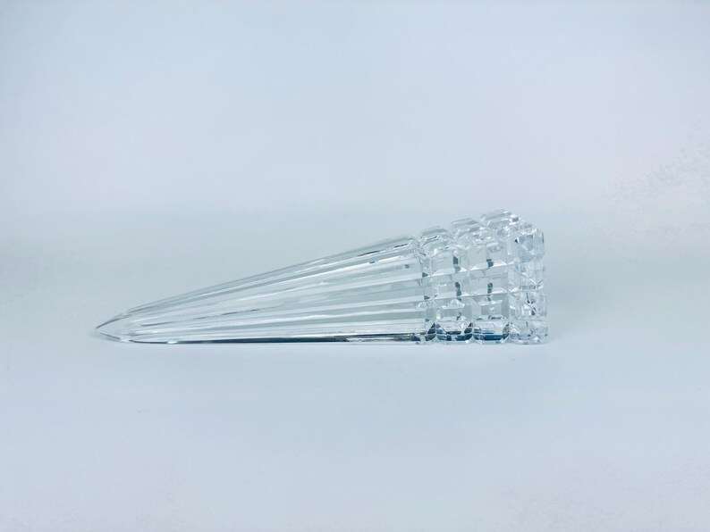 May include: A clear, glass door stop with a geometric, textured end and a tapered, ridged body. The door stop is lying horizontally on a white surface. The design is modern and minimalist.