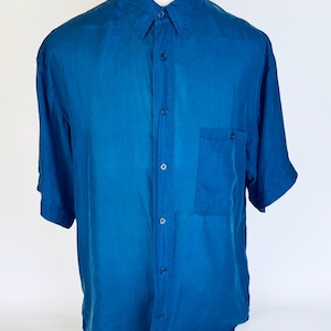 以下が含まれることがあります： 白いマネキンに展示された、胸ポケット付きのティール色の半袖シャツ。生地はわずかに光沢があります。シャツはボタンダウンカラーで、クラシックでカジュアルなスタイルです。シャツは鮮やかな色合いです。