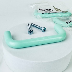 Peut inclure: Une poignée de placard en plastique vert clair avec deux vis argentées. La poignée est sur une surface blanche. L'emballage de la poignée est visible en arrière-plan.