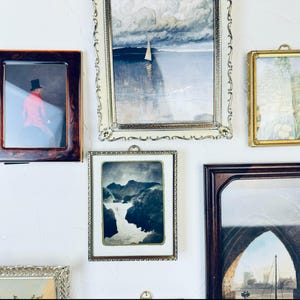 May include: A collection of framed art pieces, including landscape and portrait paintings, displayed on a white wall. Frames vary in style, from ornate gold to simple wood, showcasing diverse artistic styles and periods. The arrangement creates a gallery-like aesthetic.