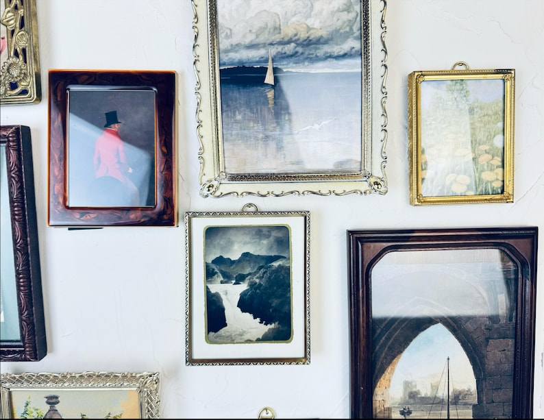 May include: A collection of framed art pieces, including landscape and portrait paintings, displayed on a white wall. Frames vary in style, from ornate gold to simple wood, showcasing diverse artistic styles and periods. The arrangement creates a gallery-like aesthetic.