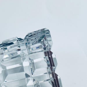 May include: Close-up of a clear, rectangular crystal object with a geometric, faceted design. The object has a repeating pattern of square and rectangular shapes, reflecting light. The background is a plain white.