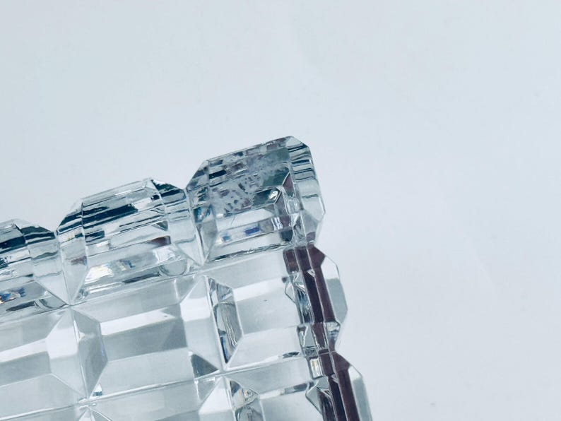 May include: Close-up of a clear, rectangular crystal object with a geometric, faceted design. The object has a repeating pattern of square and rectangular shapes, reflecting light. The background is a plain white.