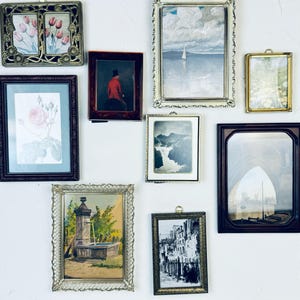 May include: A collection of framed art pieces, including floral prints, landscapes, and a portrait of a person in a red coat. The frames vary in style, from ornate gold to simple dark wood, creating a gallery wall aesthetic.