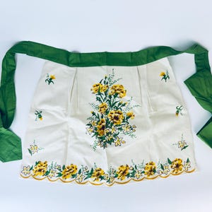 Puede incluir: Un delantal blanco con un lazo verde y un estampado floral de flores amarillas y hojas verdes. El delantal tiene un borde festoneado.
